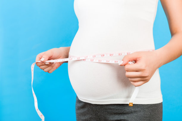 在蓝色背景下，孕妇穿着家装，用卷尺检查她日益增长的肚子。寸测量。本空间图片下载