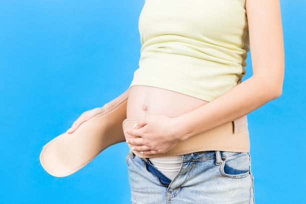 蓝色背景下孕妇在腹部系上孕妇腰带的特写镜头。骨科腹部支撑带概念。本空间图片下载