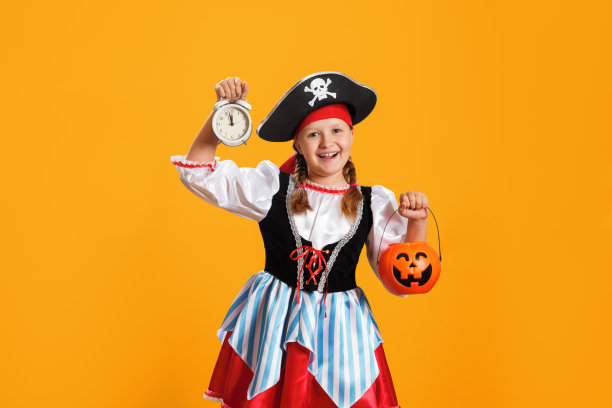 万圣节。在画室里的黄色背景上画一个穿着狂欢节服装的海盗小女孩。孩子拿着一个闹钟和一个南瓜灯笼形状的水桶。图片下载