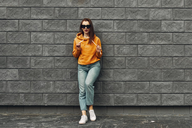 夏天的背景。穿着橙色连帽衫的漂亮女孩，靠在灰色的墙上。图片下载