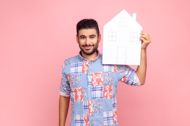 有魅力的男人留着胡须，穿着蓝色休闲衬衫，拿着纸房子，看着镜头，面带微笑，梦想着自己的家图片下载