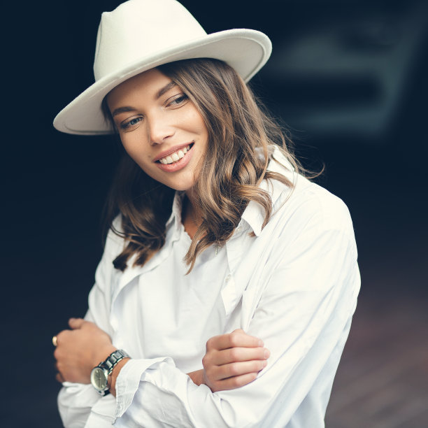 穿着白色裤子和白色棉质衬衫的时尚女孩靠墙摆姿势。一个戴帽子的女人的照片。图片下载