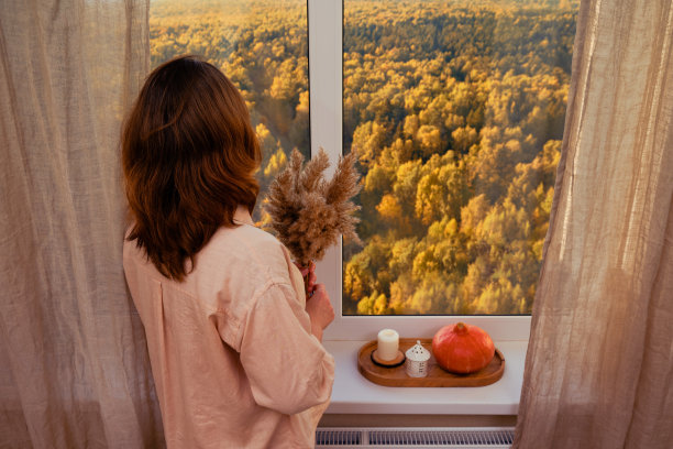 一个穿着粉红色衬衫的女人看着站在窗前的秋天的树图片下载