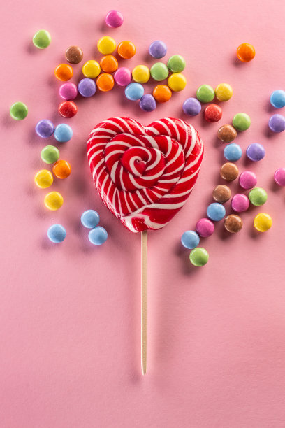 心形的条纹红色棒棒糖周围的甜蜜彩色糖果躺在粉红色的背景。图片下载
