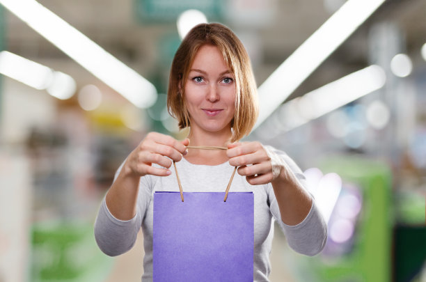 购物的概念。一个金发女郎的肖像，双手拿着一个紫色的纸板袋。背景是模糊的商店交易大厅图片下载