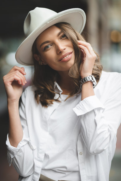 穿着白色裤子和白色棉质衬衫的时尚女孩靠墙摆姿势。一个戴帽子的女人的照片。图片下载