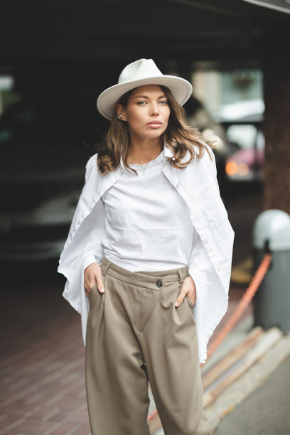 穿着白色裤子和白色棉质衬衫的时尚女孩靠墙摆姿势。一个戴帽子的女人的照片。图片下载