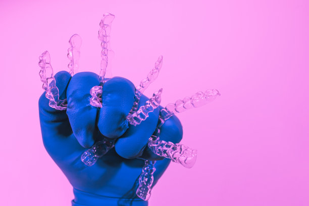 在粉红色背景下，一只戴着蓝色手套的手拿着隐形的牙套。牙科用塑料牙套矫正牙齿。图片下载