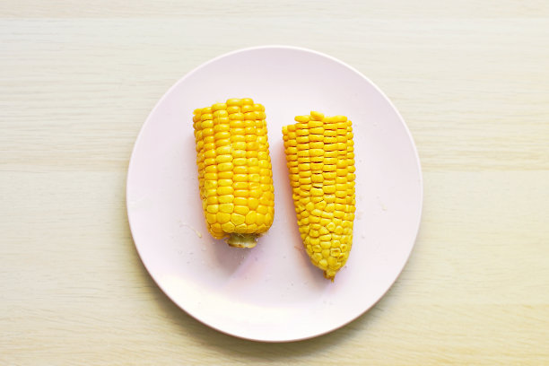 一穗热煮黄玉米被掰成两半，躺在棕色桌子上粉红色的圆形平板上，俯视图，复制空间。图片下载