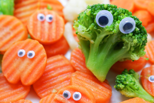 全帧图像的冷冻混合蔬菜堆在一个白色的盘子上，瞪大的眼睛添加了皱纹切胡萝卜片和花椰菜和花椰菜的小花，儿童健康饮食，幽默的概念图片下载