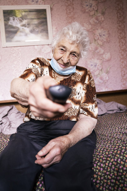 一位祖母在电视上换频道。在COVID-19期间戴着防护口罩看电视的老年妇女。一位女性的双手用遥控器布满皱纹的特写。图片下载