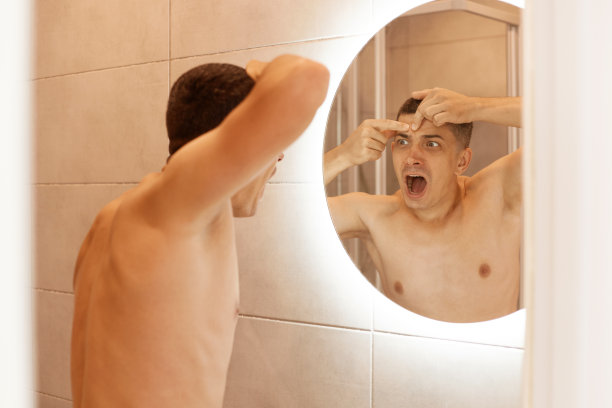 在室内拍摄的照片中，一个年轻的黑发男子对着镜子尖叫，因为它发现了皮肤上的问题，发现了额头上的痤疮，嘴巴张得大大的。图片下载