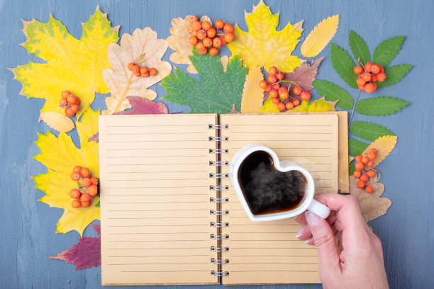 一只手拿着一个白色的心形杯子，里面有热气腾腾的咖啡或茶，还有一个空白的有线条的手工记事本图片下载