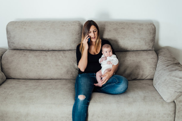 一位母亲坐在家里的沙发上打电话，怀里抱着她的孩子。图片下载