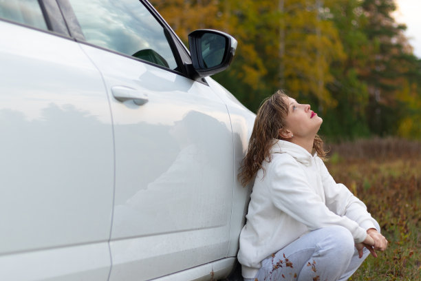 一个穿着白色连帽衫和运动裤的漂亮年轻女子坐在一辆白色汽车旁边，在一个温暖的秋日里，映衬着黄色树叶的背景。有选择性的重点图片下载