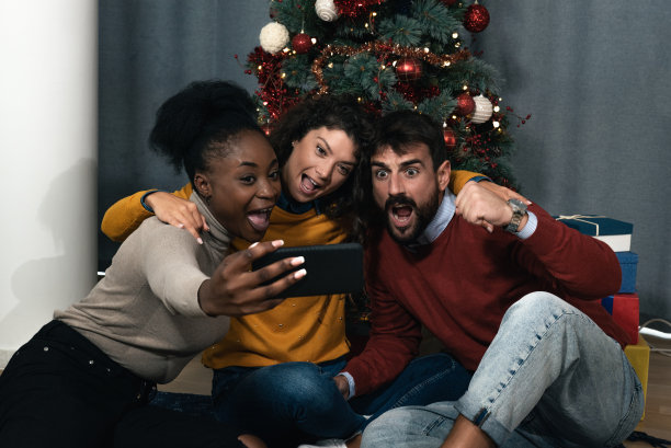 三个年轻的朋友一起庆祝圣诞节和新年前夜，在圣诞树前开玩笑，用智能手机自拍，做出有趣的面部表情图片下载