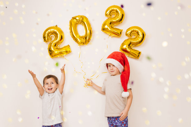 孩子们戴着红色的圣诞帽，开心地微笑着，在挂着金色气球的白色墙壁旁，享受着五彩纸屑。在家里庆祝2022年新年。圣诞节的概念图片下载