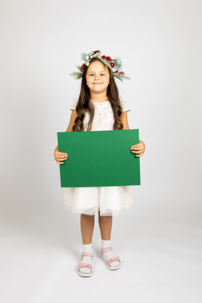 可爱的学龄前女孩的全身肖像，戴着圣诞花环，穿着白色的节日礼服，手里拿着绿色的空海报，孤立在白色背景上图片下载
