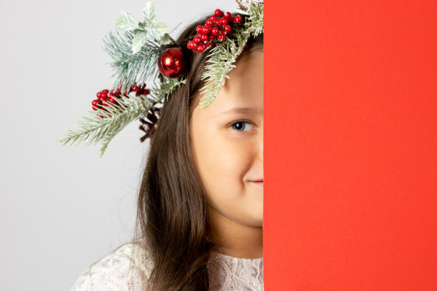特写的六岁白人女孩在圣诞花环隐藏半脸背后的红色空海报，孤立在白色背景图片下载