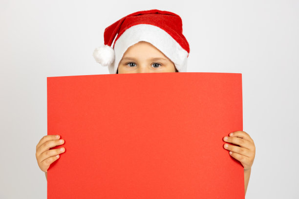 可爱的孩子的特写肖像戴着圣诞老人的帽子躲在红色的空海报后面，在白色的背景上孤立图片下载