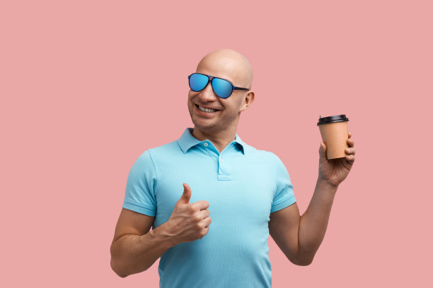 早上好。开朗微笑的光头男子享受他的热咖啡，竖起大拇指，拿着外卖杯，友好，穿着蓝色polo衫和太阳镜，站在粉红色的背景上。图片下载