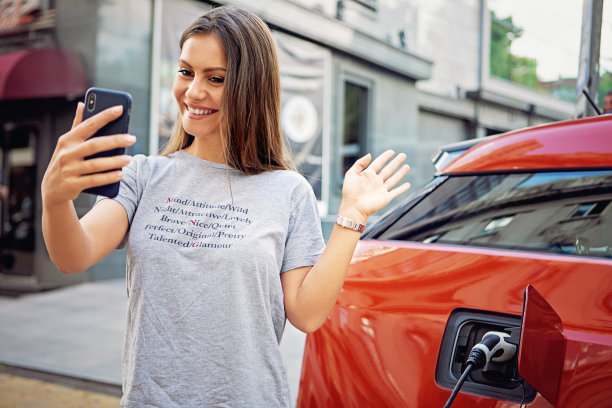 一名年轻女子正在自拍/打视频电话，直到给她的电动汽车充电图片下载