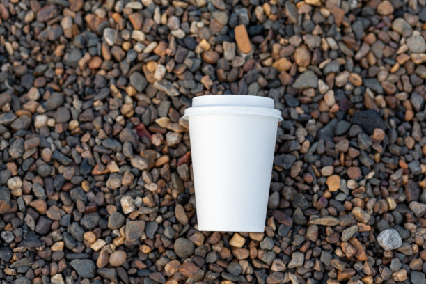 一个白色的纸杯咖啡或茶躺在岩石岸边，卵石图片下载
