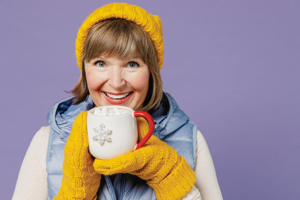 成熟的老人年长的女士女人55岁穿着蓝色马甲黄色帽子手套看相机拿喝杯咖啡与棉花糖孤立素色蜡笔淡紫色背景摄影棚肖像图片下载