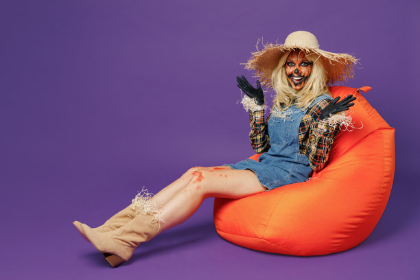 全身有趣的年轻女子20多岁，带着万圣节化妆面具，戴着草帽，穿着稻草人服装，坐在包椅上，双手分开，在纯暗紫色背景的工作室庆祝节日派对概念。图片下载