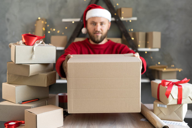 圣诞老人手里拿着礼物的盒子。小企业和圣诞快递。戴着圣诞老人帽子的大胡子年轻人正在包装礼物。邮寄度假。商业和创业图片下载