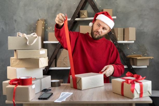 一名戴着圣诞帽的男子在仓库里准备圣诞礼物并系上红丝带。桌子上有纸板箱。新的一年销售图片下载