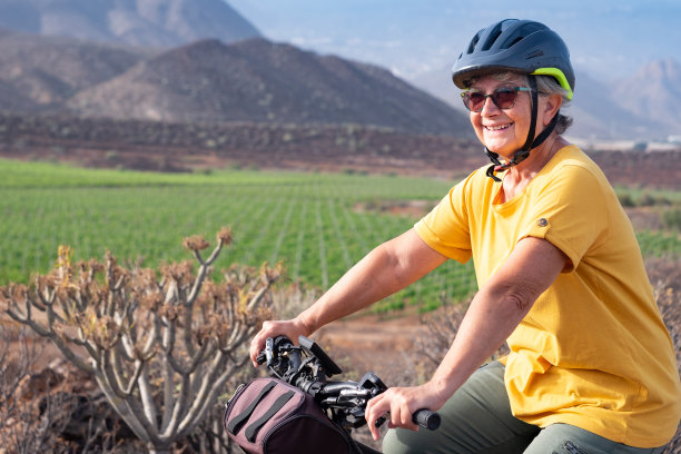 在阳光明媚的日子里，微笑的老妇人骑着她的电动自行车在户外远足，戴着运动头盔和太阳镜。绿色的葡萄园和山的背景图片下载