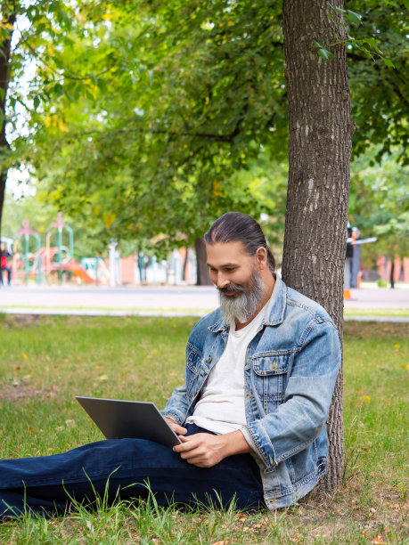 图为一名大胡子男子在用笔记本电脑上网。他坐在公园的草地上远程工作图片下载