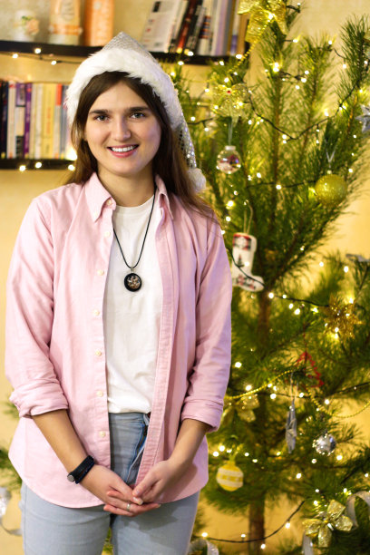 微笑的青年肖像深色女人自由职业者20-25年穿着白色圣诞老人帽和粉红色衬衫。以圣诞树为背景在室内摆造型。交叉双臂。垂直选择聚焦图片下载