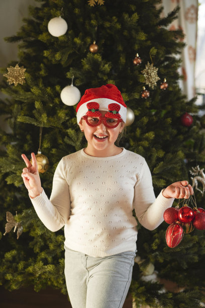 有趣的小女孩与圣诞老人的帽子和有趣的圣诞眼镜装饰圣诞树彩色球。圣诞节和新年的概念。图片下载