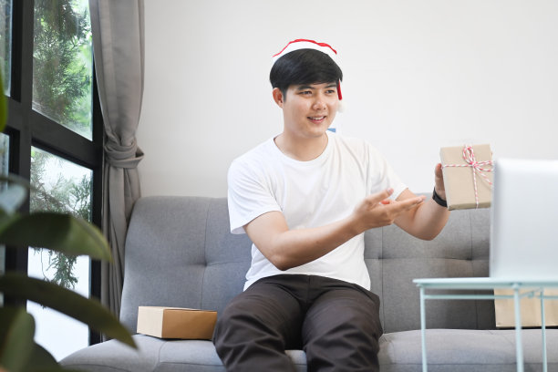 年轻人戴着圣诞帽，手里拿着礼品盒，正在家里打视频电话。图片下载