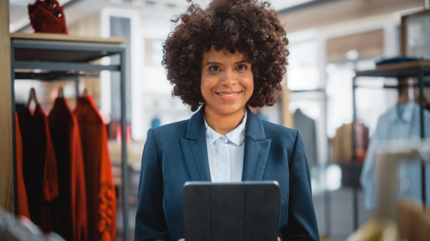 一个快乐的女性销售经理在时尚的服装店使用平板电脑的肖像。专业商店销售零售助理检查库存。小企业主订购购物中心项目。图片下载