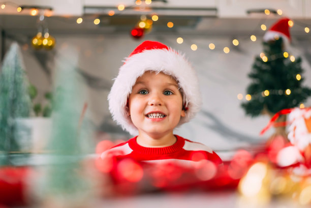有趣的蹒跚学步的男孩戴着圣诞帽坐在装饰圣诞节的厨房里。节日，传统，婴儿食品和健康概念。图片下载