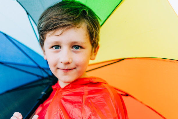 一个男生的肖像，身后撑着彩虹伞。孩子把彩色伞扛在肩上。欢快的孩子穿着红色的雨衣，撑着五彩的雨伞图片下载