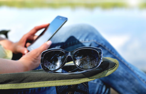 太阳眼镜躺在躺椅的扶手上，女孩在扶手椅上看着手机，背景是大自然，河边是绿色的夏天夏天阳光灿烂的日子特写图片下载