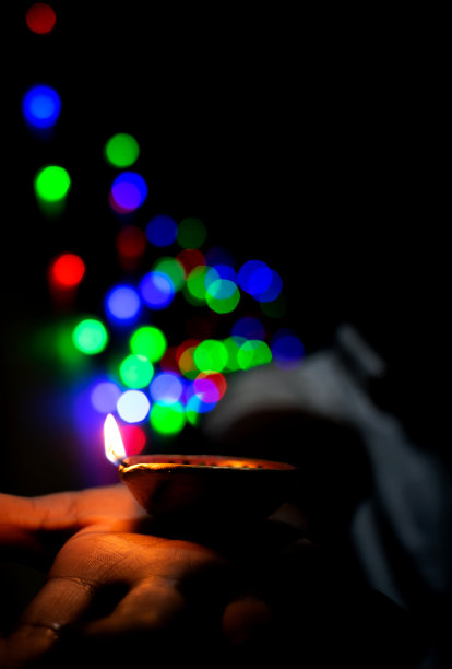 照亮了排灯节diya由地球上的男孩的手与选择性聚焦和模糊的彩色散景灯背景在晚上。图片下载