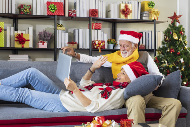 白种老人夫妇在圣诞树装饰的房间里，戴着红色圣诞老人帽，用笔记本电脑与家人视频通话，一起快乐地庆祝圣诞节图片下载