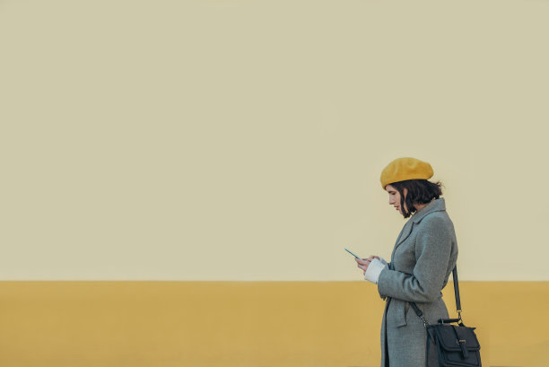戴着黄色贝雷帽和灰色外套的女人在用智能手机图片下载