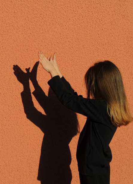 可爱的小女孩在橘色的背景下玩影子。穿黑衣服的孩子在户外投下阴影。图片下载