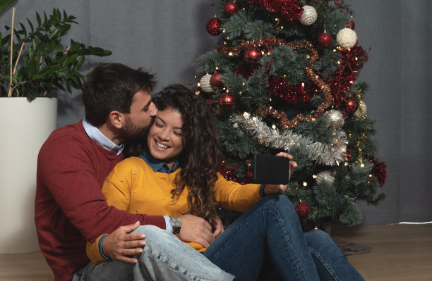 年轻幸福的情侣们坐在家里装饰着装饰品的圣诞树前自拍，期待着新年的喜庆心情，圣诞节的概念图片下载