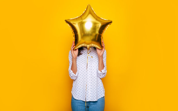 一个小女孩用黄色背景上孤立的星星形状的气球遮住了她的脸，这是一张特写照片图片下载