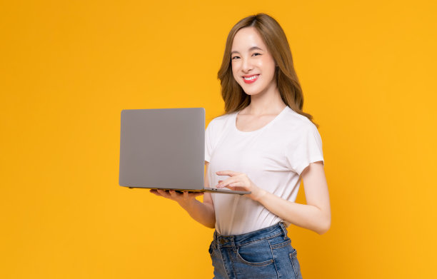 摄影室拍摄的开朗美丽的亚洲女人在白色t恤和拿着笔记本电脑打字键盘上的黄色背景。图片下载