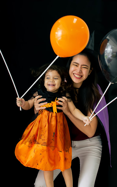 可爱的小女孩肖像，美丽的妈妈穿着女巫服装，幸福的微笑，乐趣，玩气球一起庆祝万圣节派对在黑色孤立的背景剪纸。图片下载