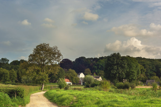 弯弯曲曲的土路穿过长满树木和房屋的草地，在蓝色多云的天空下，夏日连绵起伏的景色中。图片下载