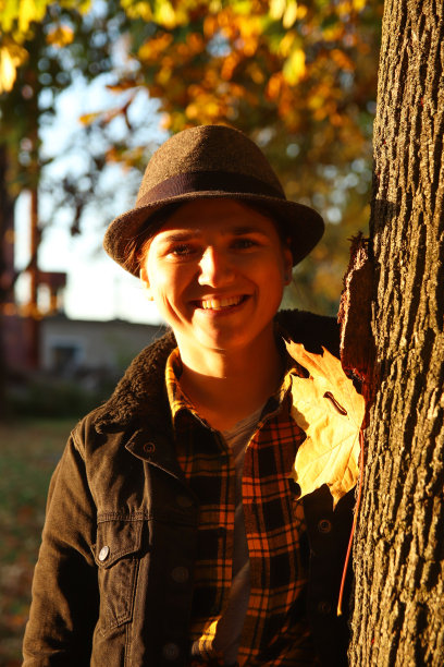 特写照片的微笑年轻的白人妇女在五颜六色的橘色秋天公园。明亮时尚的女人在帽子和牛仔裤夹克附近的树干。你好,秋天的概念。黄色的枫叶。热爱自然图片下载